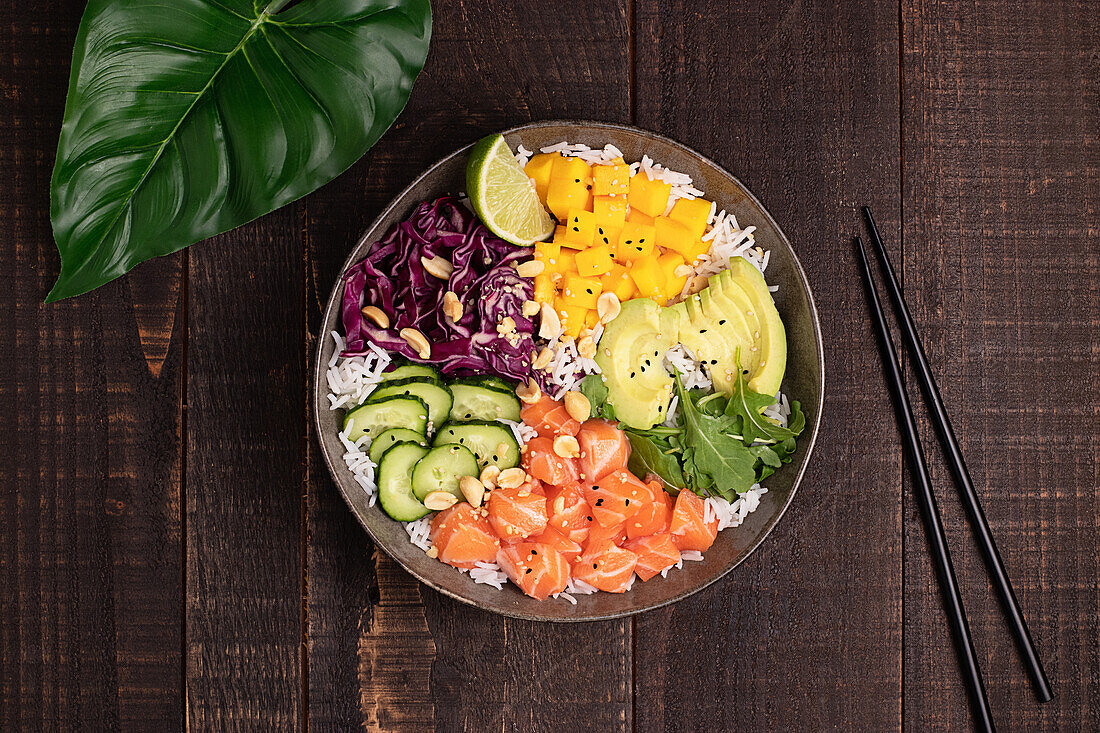 Top view of appetizing colorful poke bowl with fresh salmon and rice served with ripe avocado slices, cabbage and mango placed on wooden tray