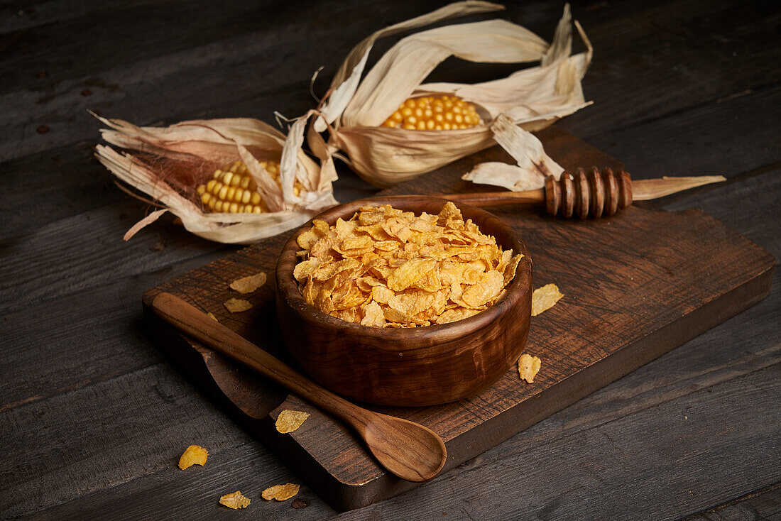 Schüssel mit leckeren süßen Cornflakes mit Löffel auf hölzernen Schneidebrett auf dem Tisch mit Maiskolben in hellen Küche platziert