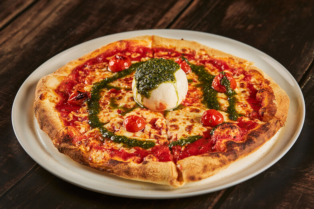 Leckere Pizza mit Kirschtomaten und Parmesan, garniert mit Mozzarella-Käsekugel und Pestosauce, serviert auf einem Teller auf einem Holztisch in einer Pizzeria