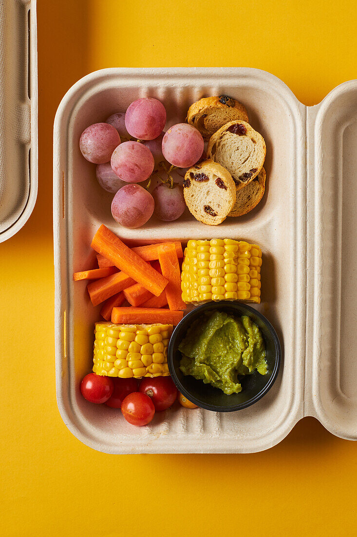 From above of lunch boxes with healthy food including crackers carrot sticks grapes cherry tomatoes with broccoli walnut and tangerine on yellow background