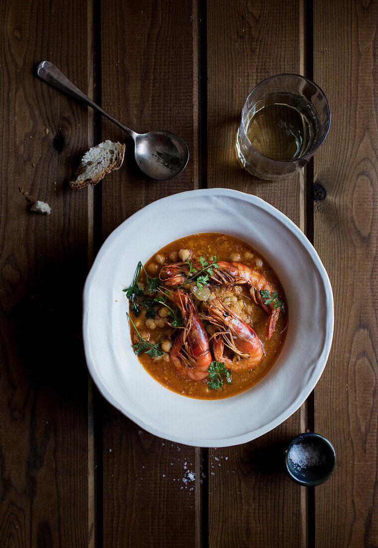 From above of delicious spicy stew with chickpeas and big shrimps decorated with cilantro leaves served in white soup plate on wooden table with glass of drink and spoon