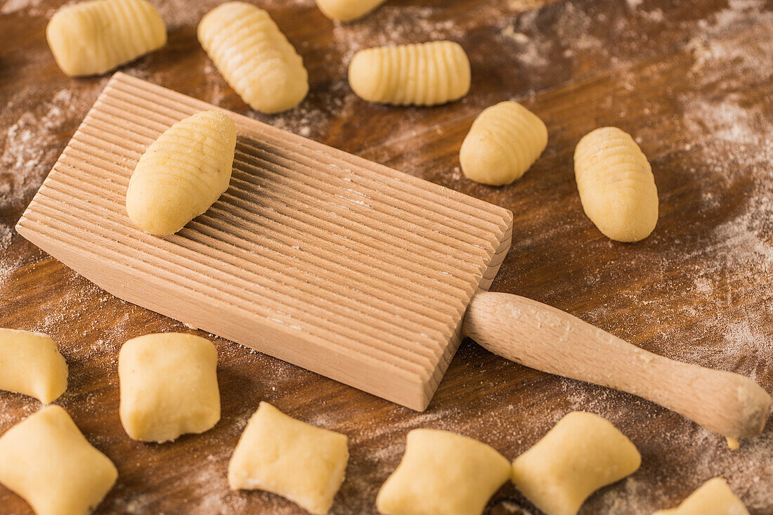 Draufsicht auf weiche, rohe Teigstücke, die während der Zubereitung von Gnocchi in der Küche auf einem mit Mehl bestäubten Holztisch neben einem Schneidebrett liegen
