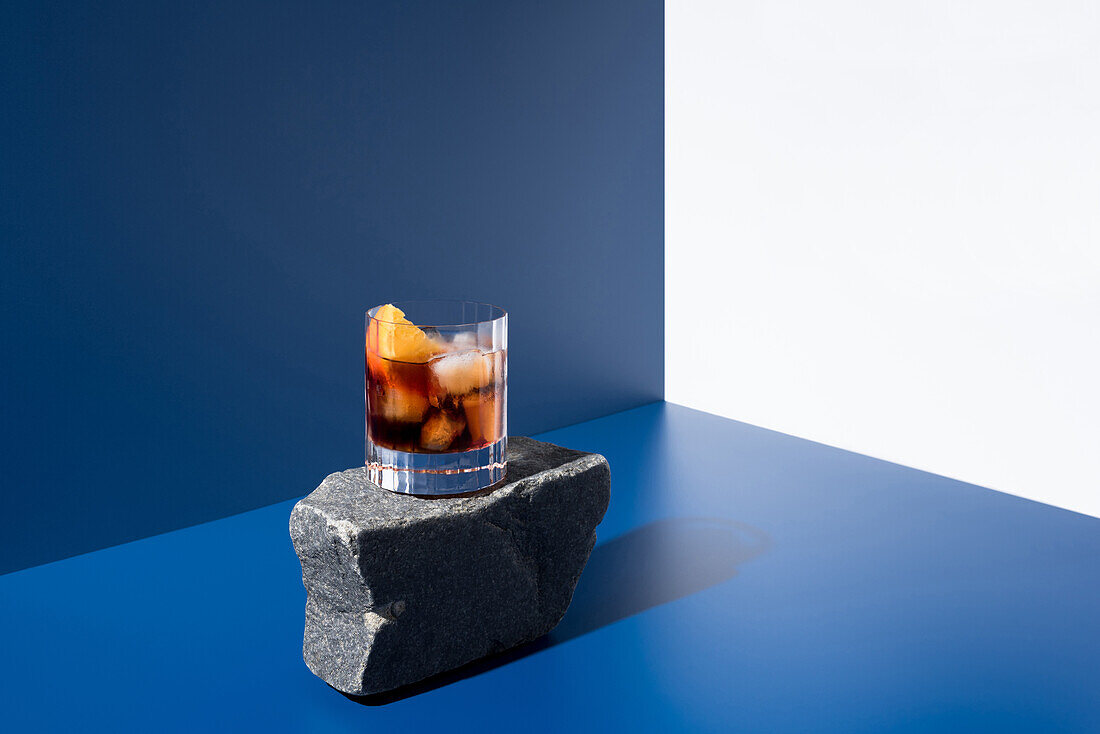 Glass of alcoholic cocktail made with red vermouth served with orange slices on stone in light studio with blue and white wall