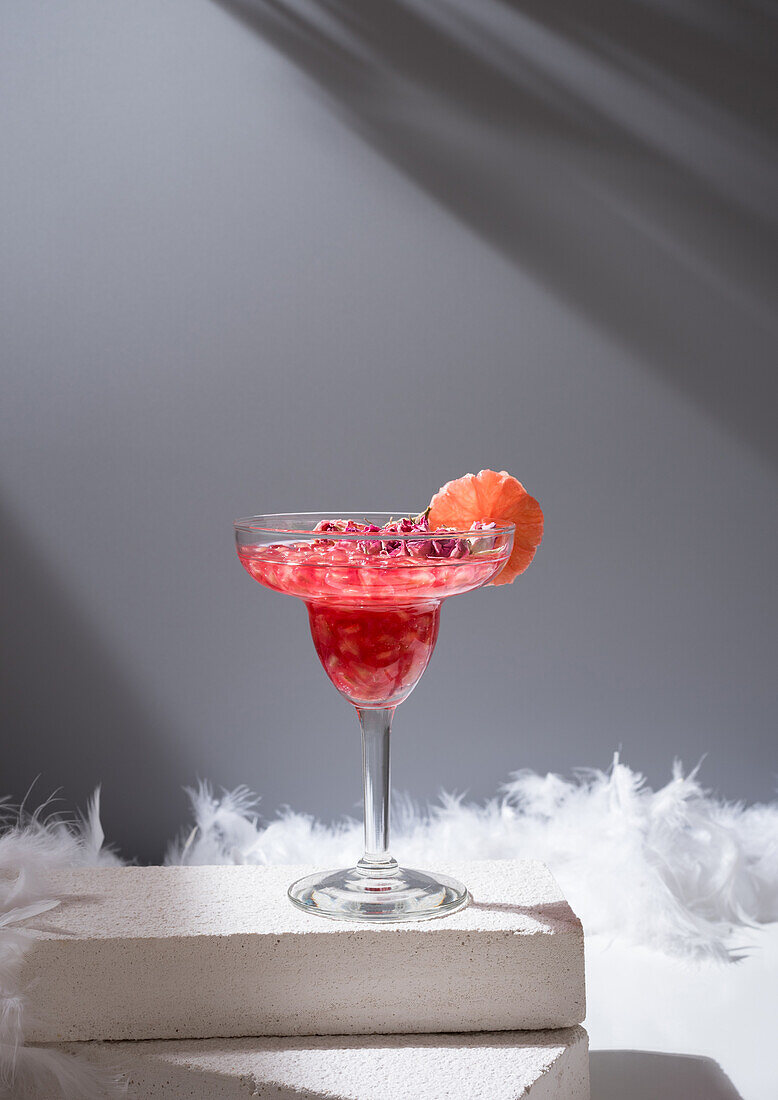 Glass of alcoholic cocktail pomegranate margarita served with slice of red orange on white blocks near feathers at sunlight