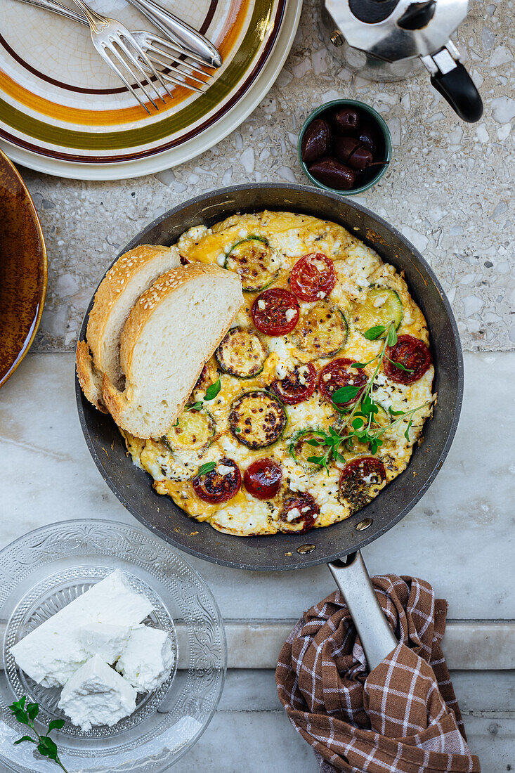 Top view of traditional Greek frittata with cherry tomatoes and feta cheese in frying pan with bread in light kitchen