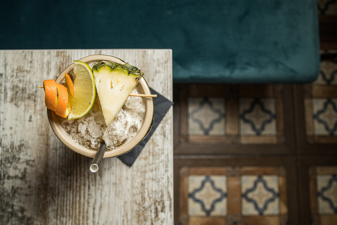 From above of large sculptural tiki cup filled with booze decorated with straw and fruits placed on green rug against wooden table