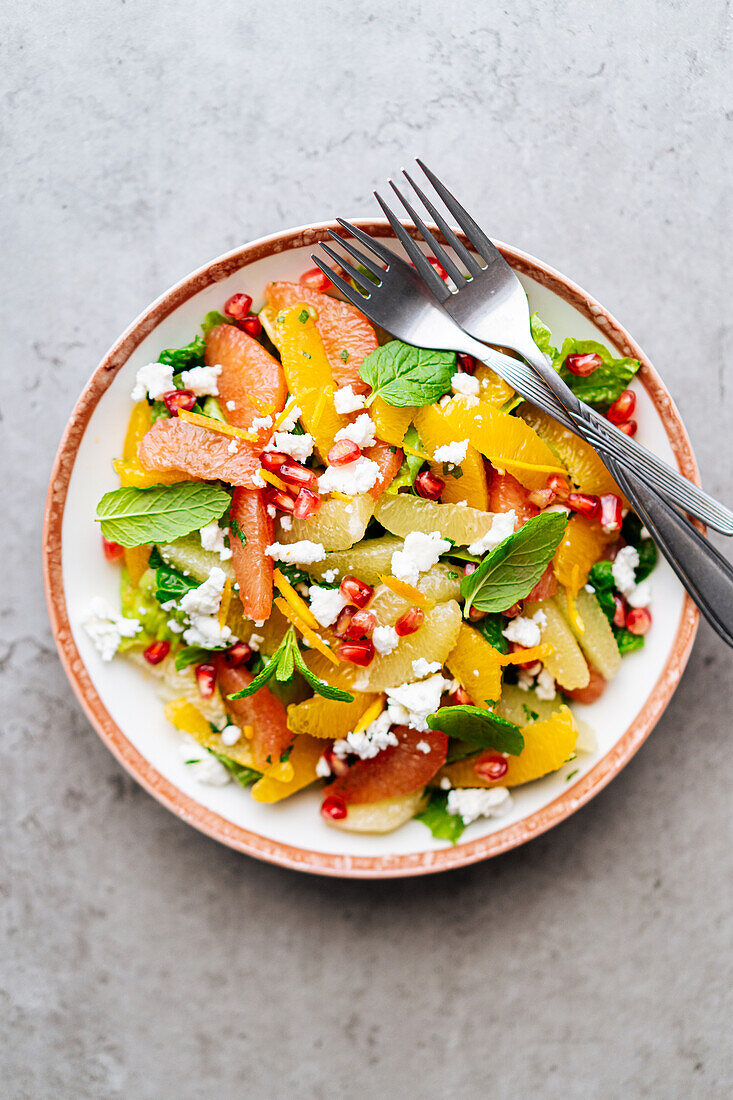 Draufsicht auf traditionellen griechischen Salat mit reifen Tomaten und Feta-Käse, gekrönt von Granatapfelkernen und Minzblättern auf grauem Hintergrund mit Orangensaft