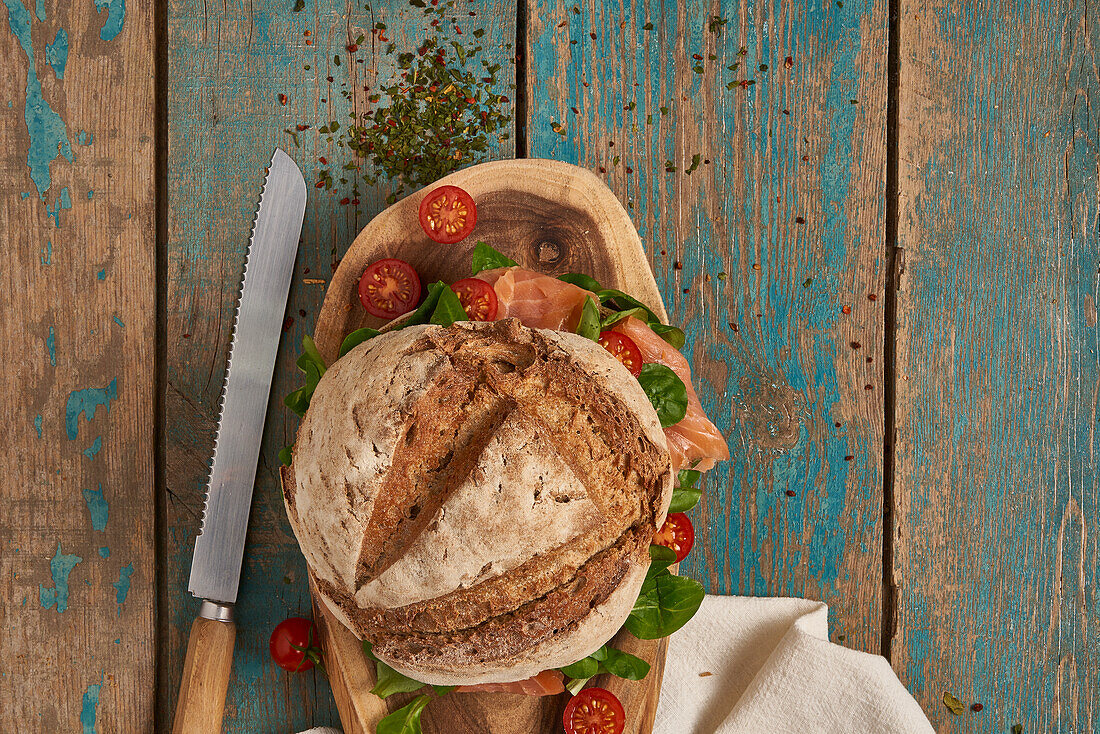 Ansicht von oben von hausgemachtem Sauerteigbrot mit Lachs, serviert auf einem hölzernen Schneidebrett mit Kirschtomaten und Messer auf einem schäbigen Tisch