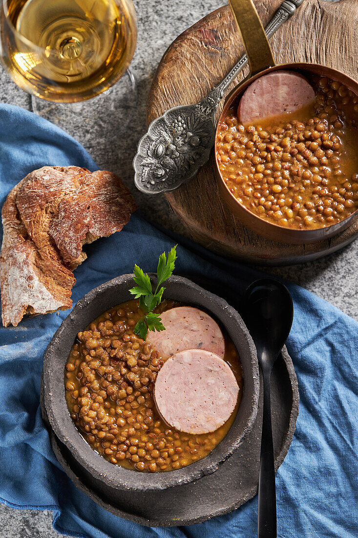 Top view of rustic bowls with tasty lentil soup with parsley and slices of sausage placed on marble table and blue napkin near bread and wine during lunch