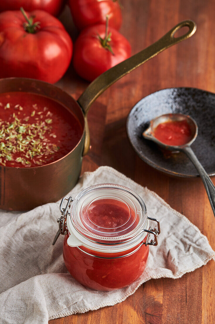 Pfanne und Behälter mit köstlichen hausgemachten Tomatensauce mit Oregano bestreut auf gewebten Matte auf Holztisch in der Küche platziert