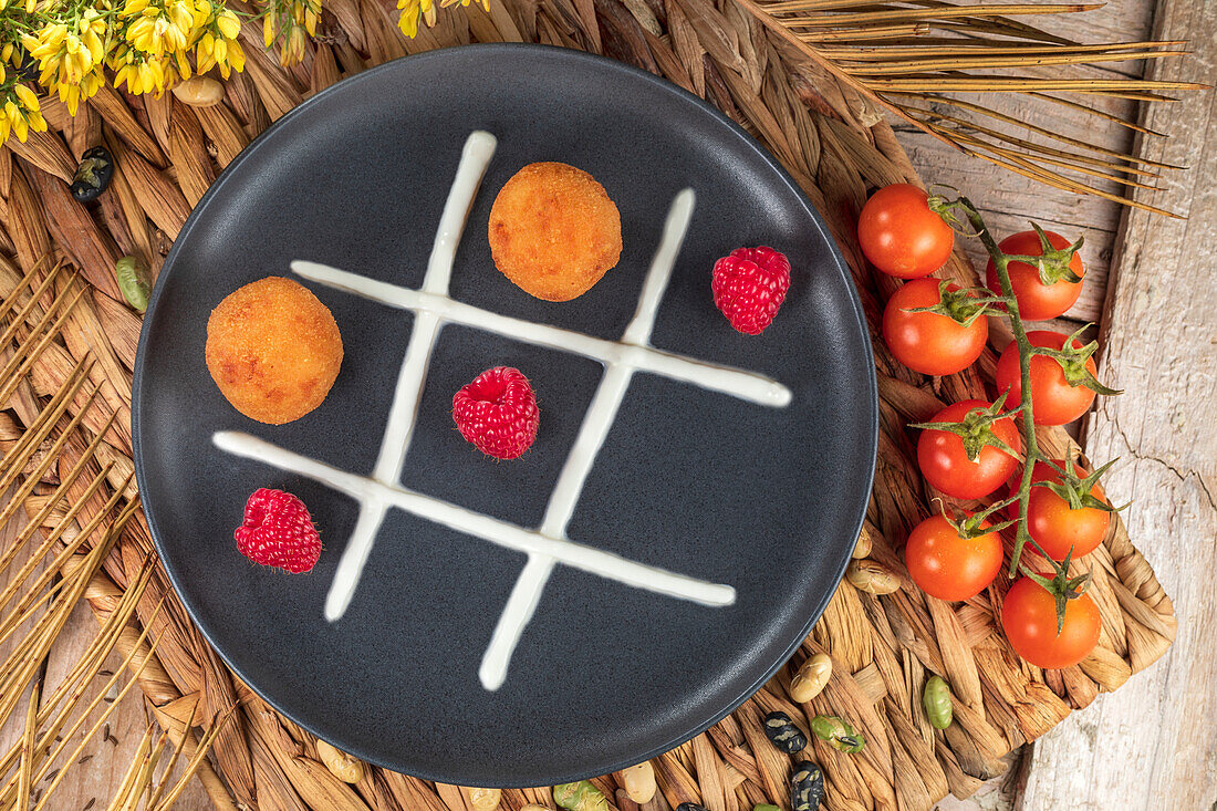Top view of tasty fritters with ripe raspberries between whipped cream representing tic tac toe game on plate near cherry tomatoes