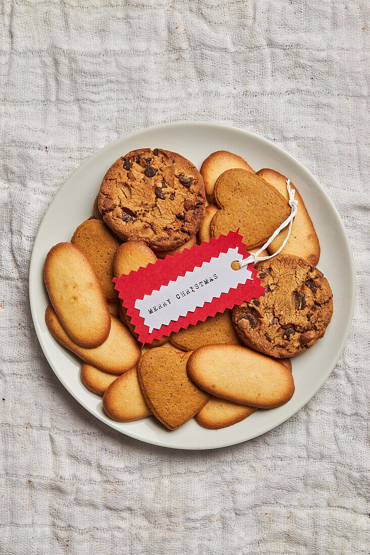 Teller mit verschiedenen süßen Keksen und Geschenkanhänger auf einem Tisch mit Weihnachtsdekoration von oben