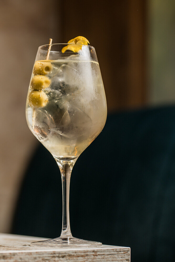 Kristallweinglas mit Martini-Cocktail, serviert mit Zitronenschale und Oliven am Rand eines Holztisches