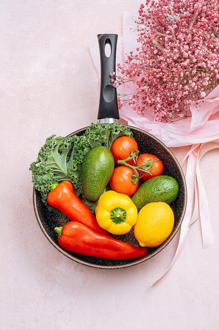 Top view of frying pan with abundance of uncooked vegetables on pink background with bouquet of Gypsophila flowers in paper wrapping