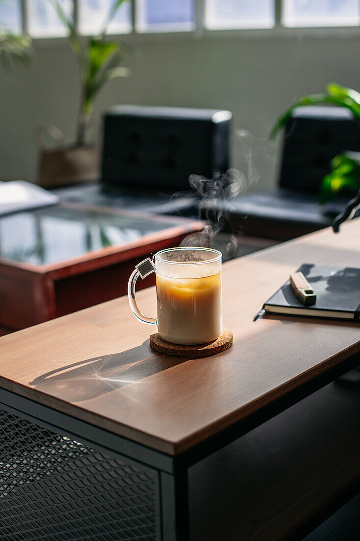 Glass of hot steaming tea with milk placed on wooden counter with notebook in sunlit apartment