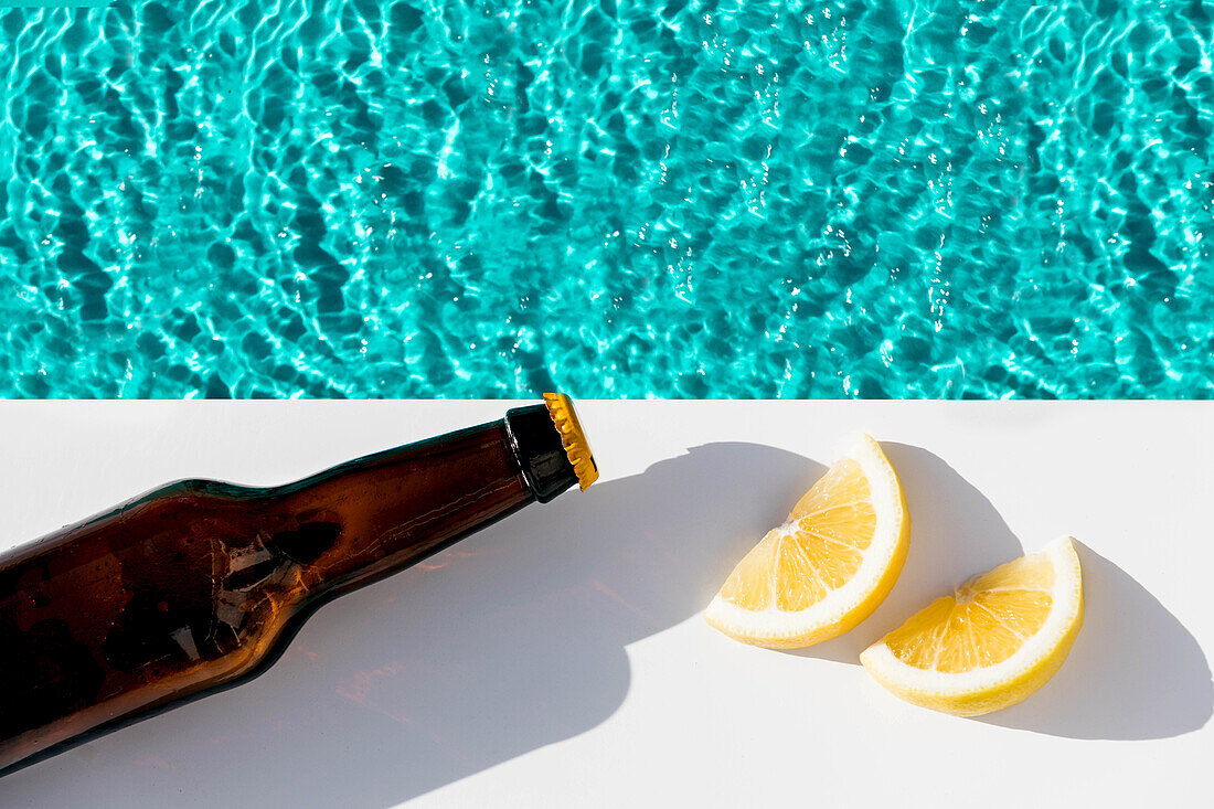 Draufsicht auf eine frisch aufgeschnittene Zitrone und eine Flasche Bier am Pool über türkisfarbenem Wasser im Sonnenlicht