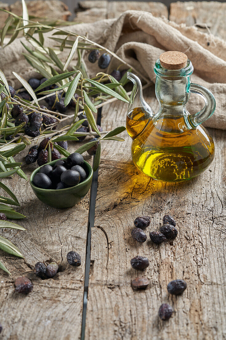 Von oben: schwarze Oliven und ein Krug mit Öl auf einem Holztisch in einer rustikalen Küche neben frischen Zweigen