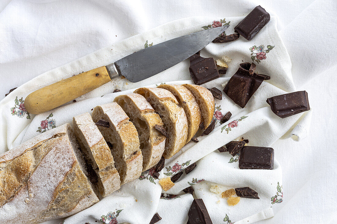High angle of freshly baked cut bread loaf with chocolate bars and knife placed on white napkin on table in kitchen