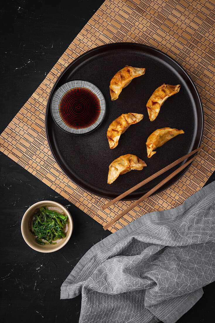 Top view of chopsticks with gyoza on plate near bowls of soy sauce and greens during lunch on black table