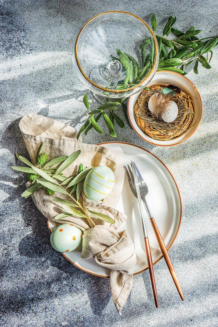 From above cutlery set for Easter dinner with olive tree branches and eggs on a bird nest on a concrete background