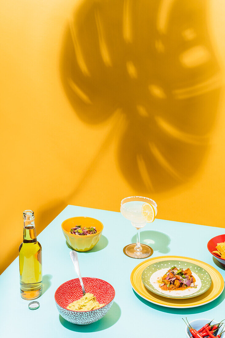 High angle of dishware with nachos and chicken salad placed near bottle of beer and glass of lemonade against yellow background