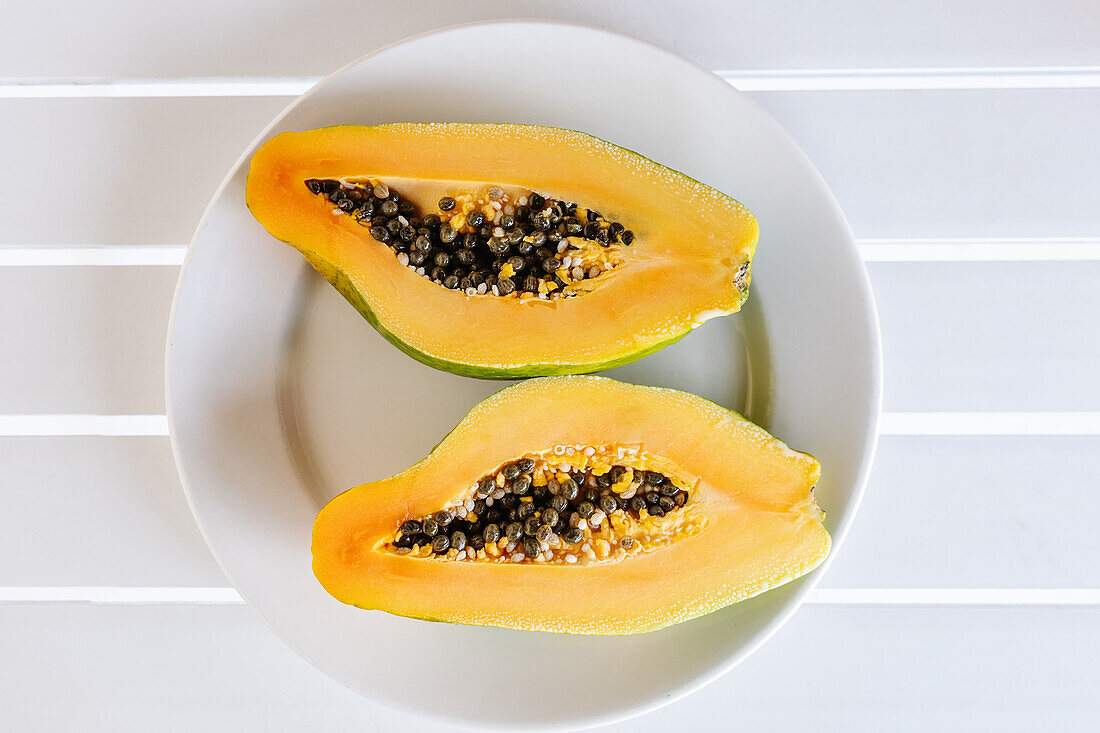 Draufsicht auf eine halbierte frische reife exotische Papaya mit Kernen, serviert auf einem Keramikteller auf einem weißen Tisch an einem sonnigen Sommertag