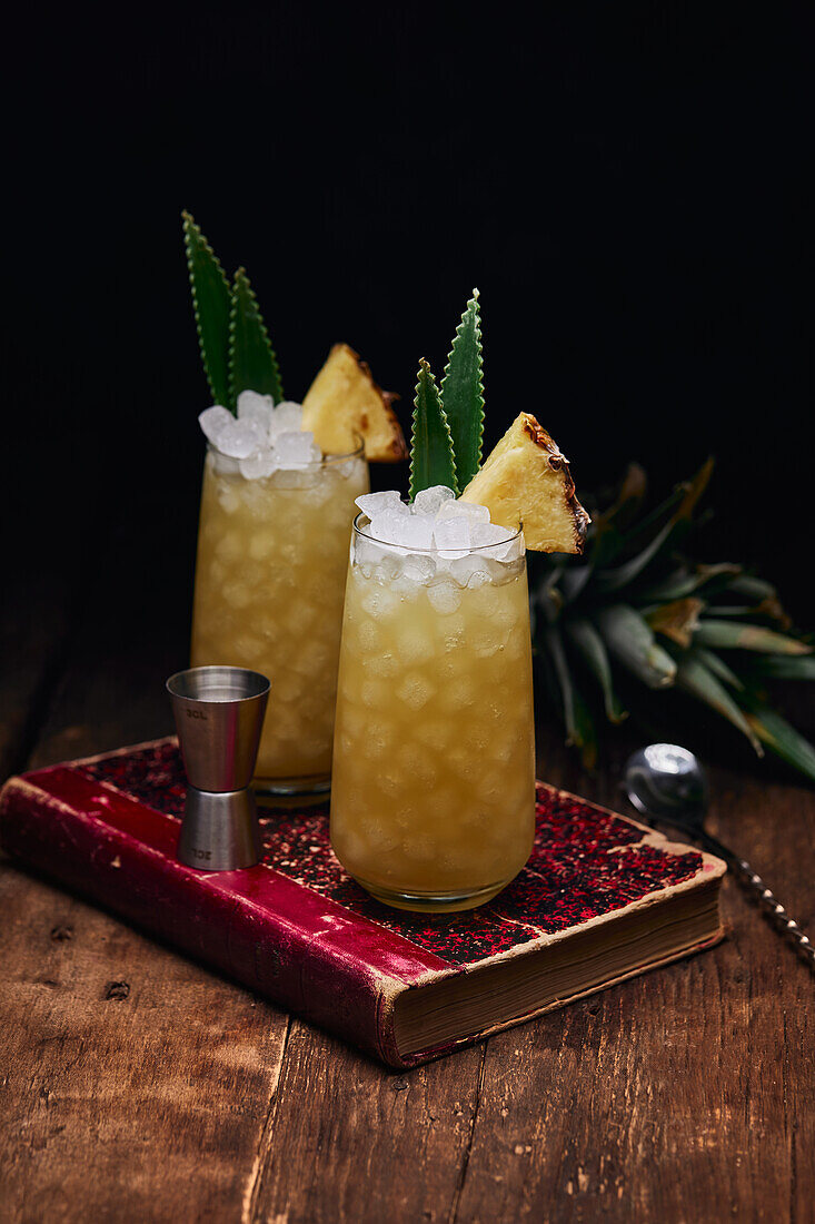 Holztisch mit Gläsern frischer gelber Cocktails mit Eiswürfeln und Ananasstücken und Blättern neben Löffel und Schnapsglas auf rotem Buch auf schwarzem Hintergrund