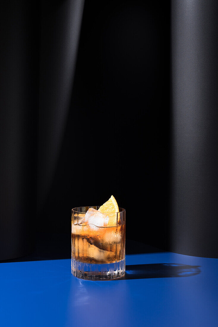 Glass of alcoholic cocktail made with red vermouth served with orange slices in light studio with blue and black wall