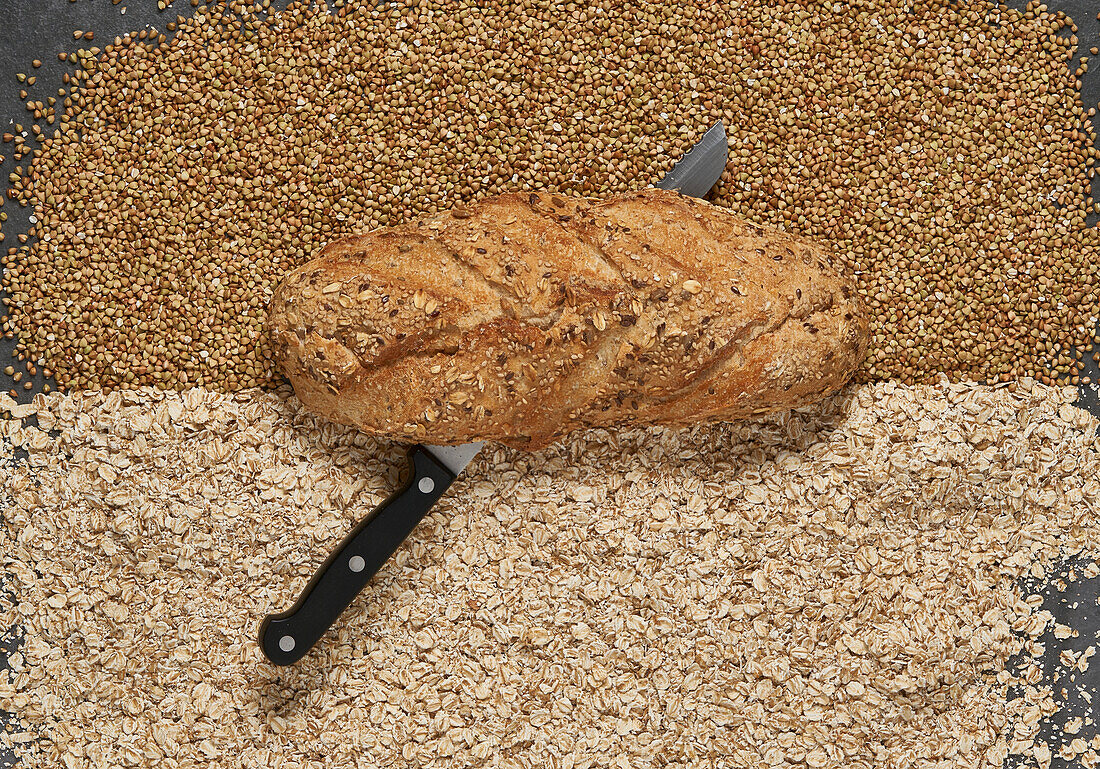 Full frame top view of freshly baked buckwheat and oatmeal bread with knife below placed on raw grains in light room