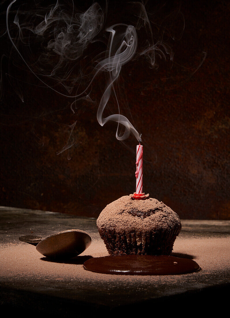Sweet birthday muffin with powdered sugar and extinguished candle served on table with spilled chocolate and spoon on dark background