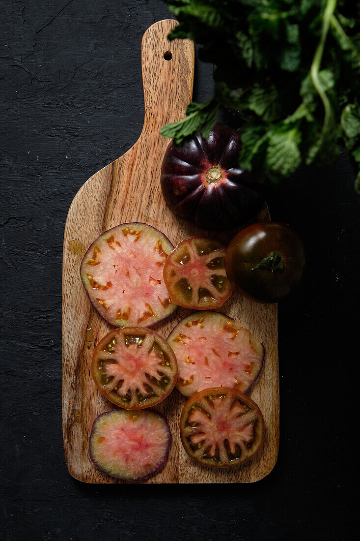 Draufsicht auf frische, reife, in Scheiben geschnittene schwarze Tomaten und grüne Minzstängel auf einem hölzernen Schneidebrett auf schwarzem Hintergrund