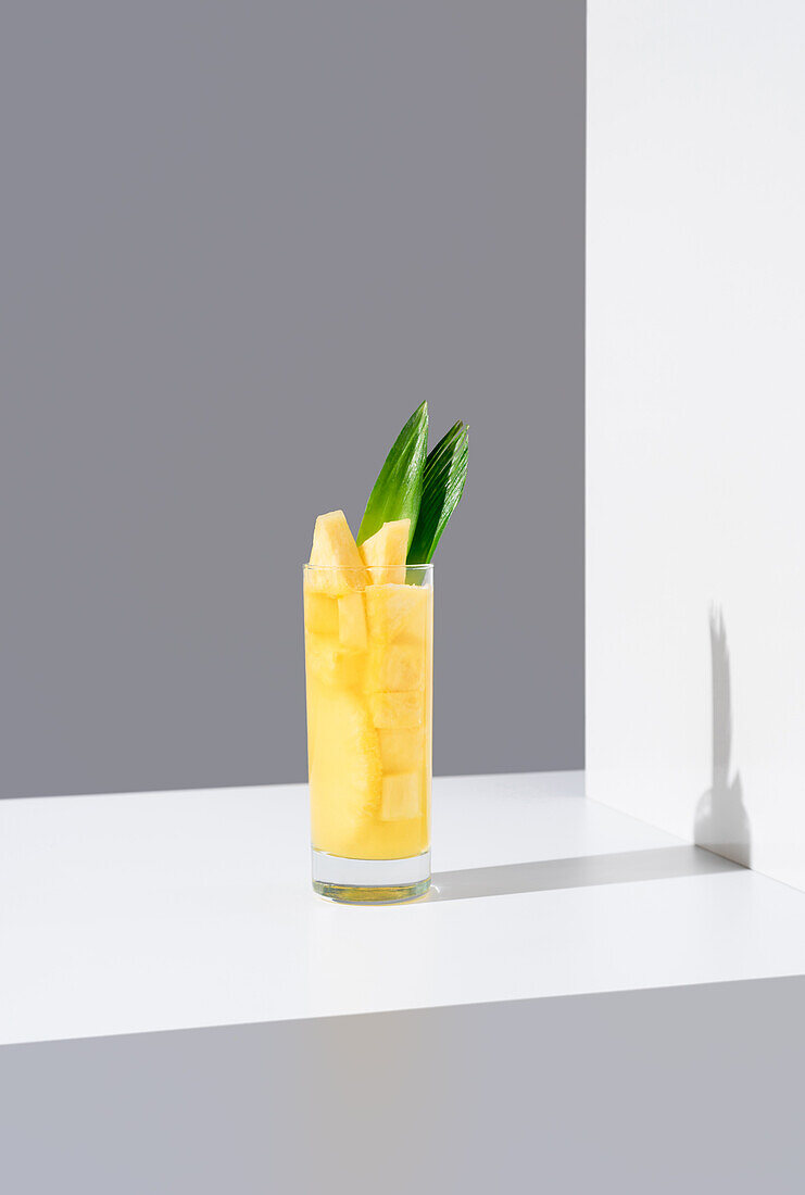 Glass full of cold pineapple juice with green leaves and straw placed on sunlit table against gray and white background