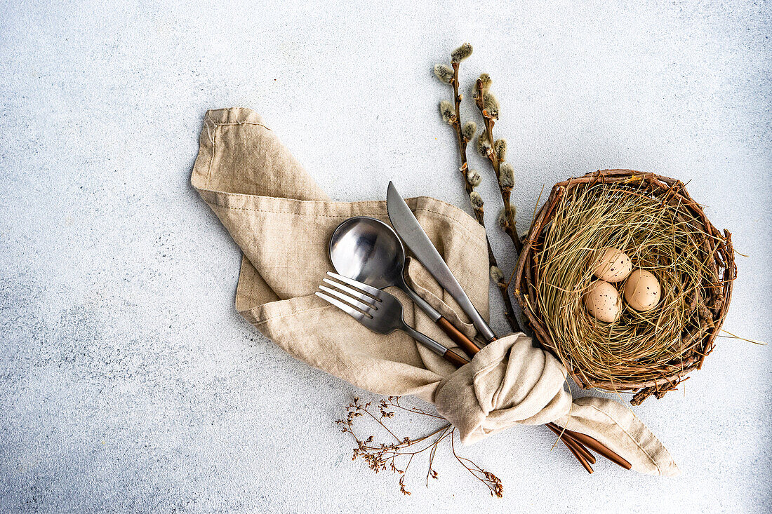 From above cutlery set for Easter dinner with dried flower twig and eggs on a bird nest on a concrete background