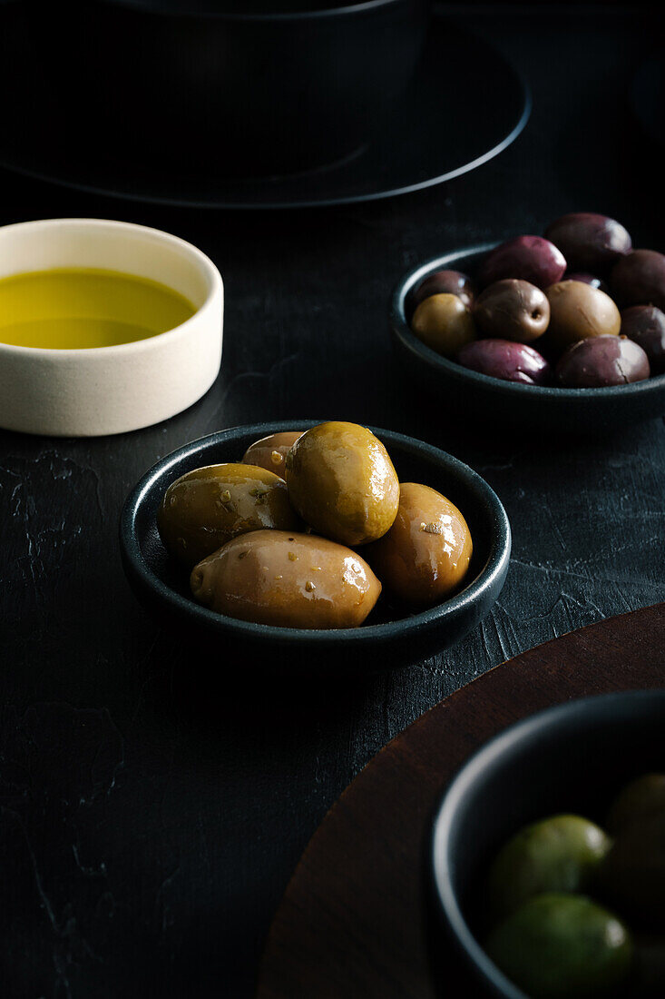 Von oben schmackhafte saftige grün-braun-gelbe Oliven und Öl in schwarz-weißen Schalen auf schwarzem Tisch
