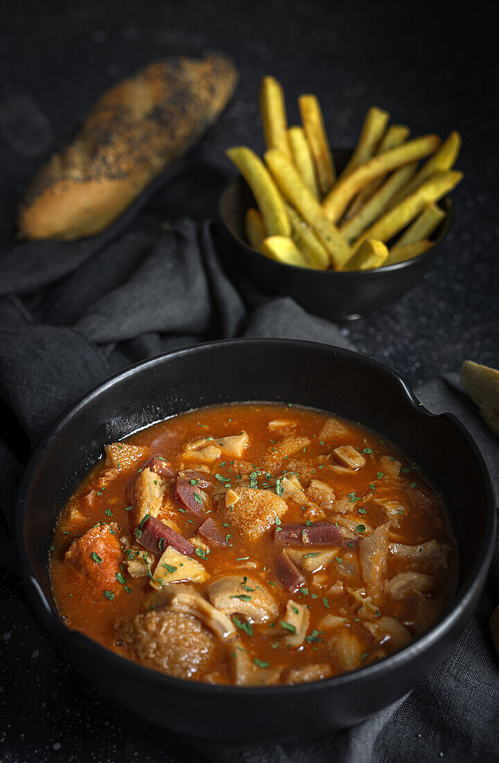 From above of tasty callos a la madrilena in black bowl near French fries and bread with seeds