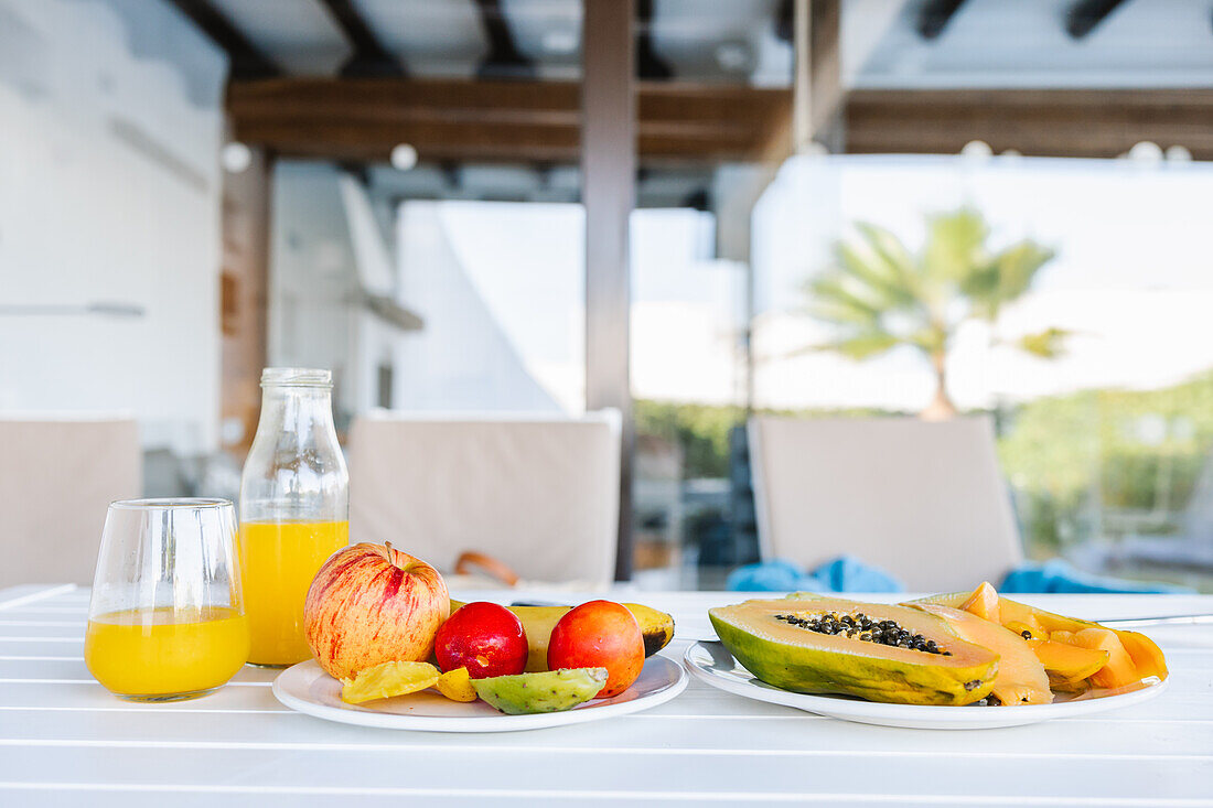 Köstliche halbierte Papaya auf einem Teller neben verschiedenen bunten Früchten und einem Glas frischen Orangensaft auf einem weißen Tisch auf der Terrasse an einem sonnigen Sommertag