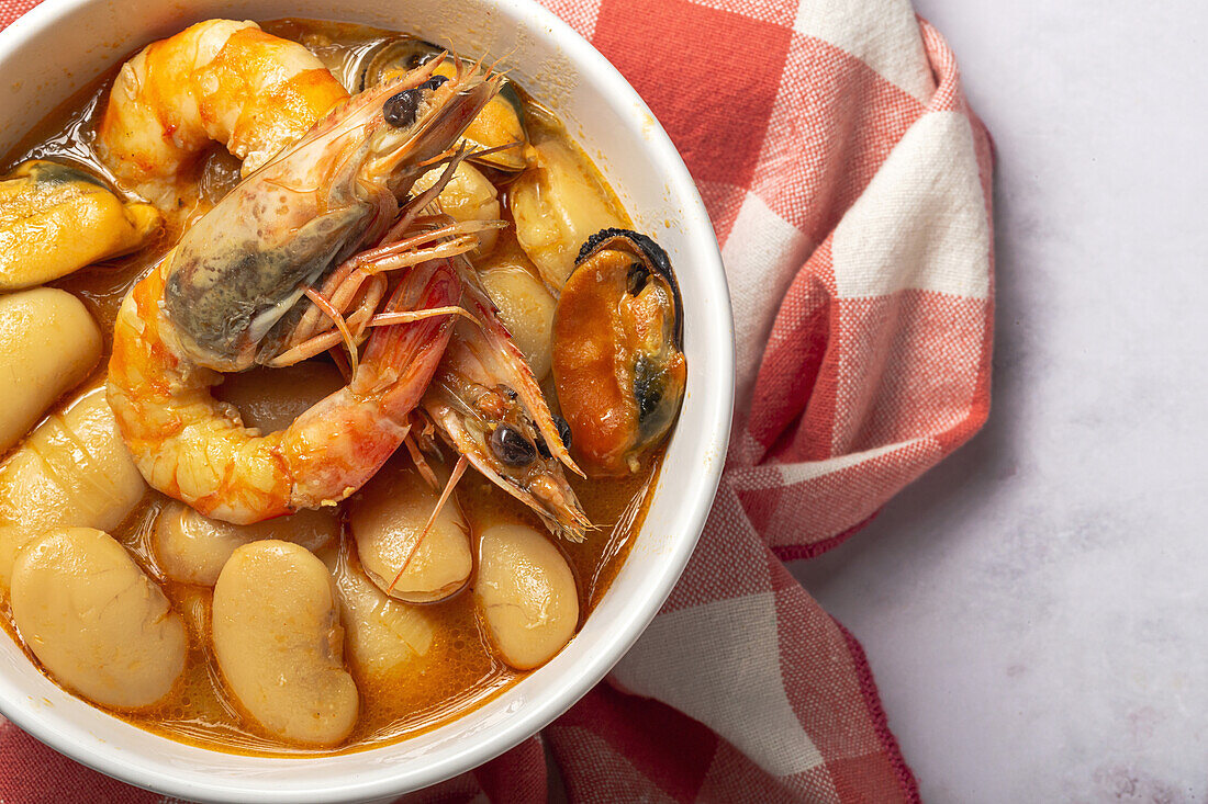 Close up of bowl with typical stew of beans with prawns, shrimps and mussels on a red checkered on a stone table