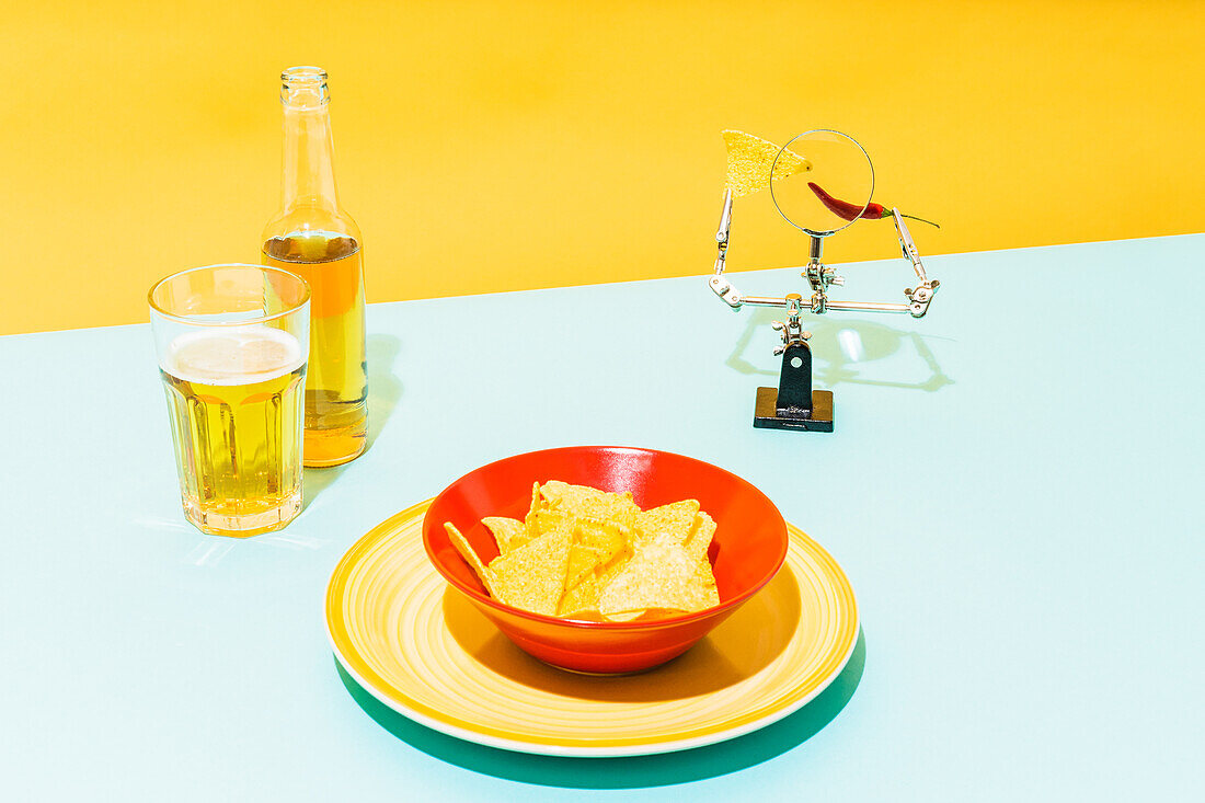 Schale mit knusprigen Tortilla-Chips neben Glas und Flasche mit kaltem Bier und Lupe mit Chilischote