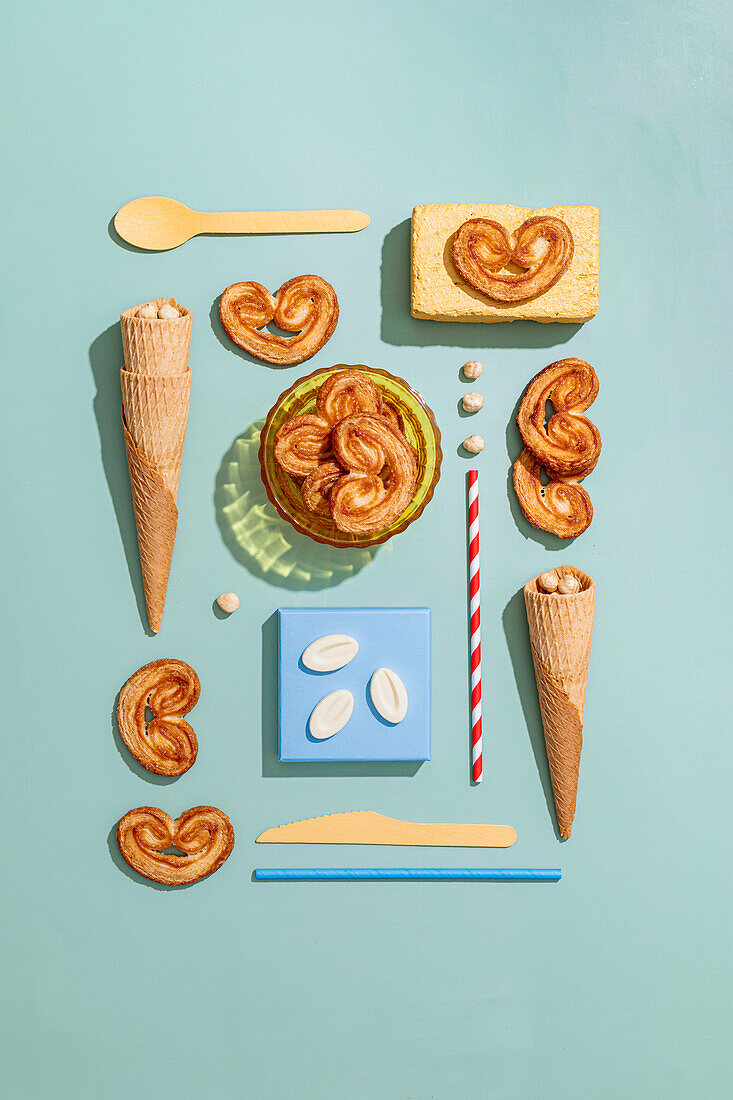 Overhead view of waffle cones and elephant ear cookies with candy placed against disposable cutlery and straw on green backdrop