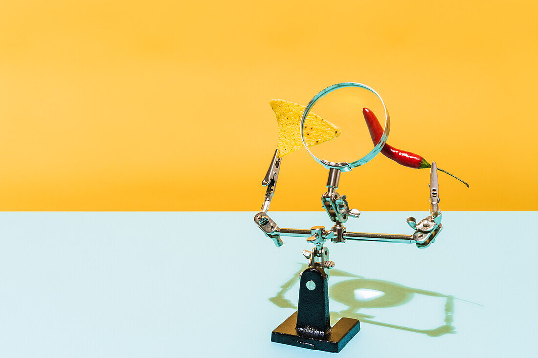 Red hot chili pepper and crunchy tortilla chip placed on mechanic holder under magnifying glass against yellow and blue background
