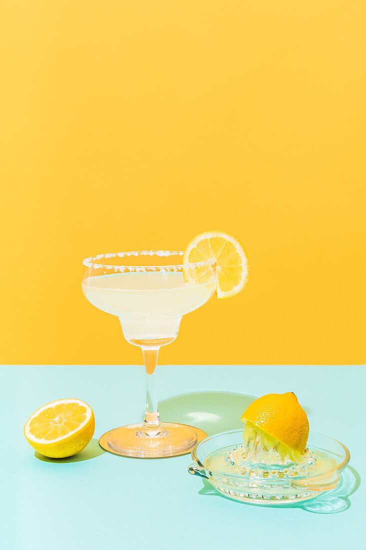 Glass and jug of cold lemonade with fresh lemons placed on colorful blue and yellow background