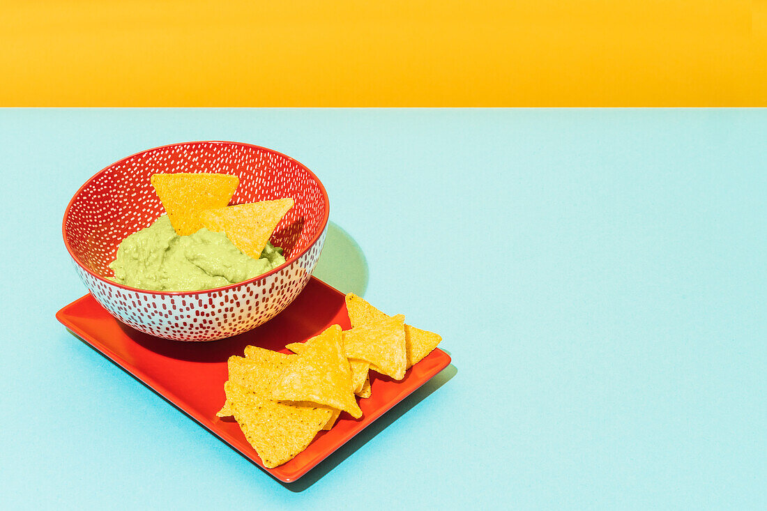 Crispy tortilla chips placed on red plate near bowl with tasty guacamole sauce on blue and yellow background