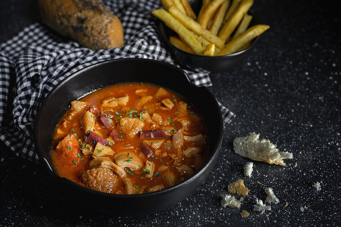 From above of tasty callos a la madrilena in black bowl near French fries and bread with seeds