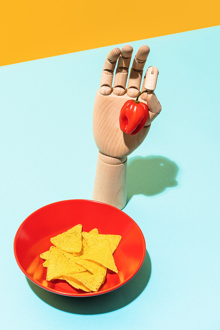 From above wooden artificial hand with red habanero pepper placed near bowl with crunchy tortilla chips of Mexican cuisine on blue and yellow background