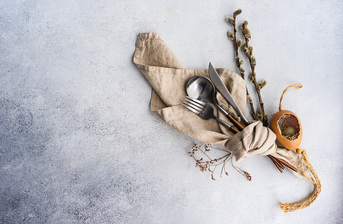 From above cutlery set for Easter dinner with dried flower twig and eggs on a bird nest on a concrete background