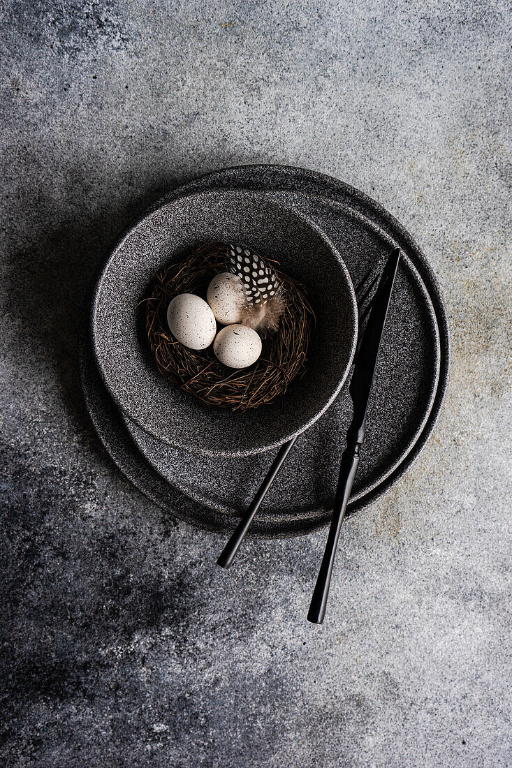 From above minimalistic table setting for Easter dinner with dark tableware and cutlery on ceramic plates and bowl with nest and quail eggs on concrete table