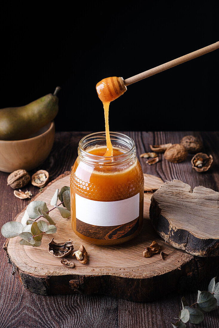 High angle trickle of natural honey flowing from wooden dipper into glass jar with blank label placed on wood slab near walnuts and pear against black background