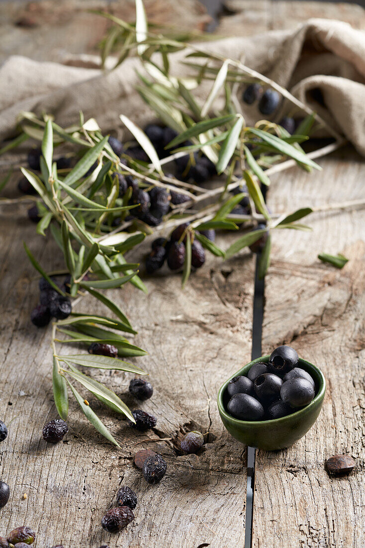 Kleine Schale mit schwarzen entkernten Oliven, die neben grünen Zweigen auf einem schäbigen Holztisch in einer rustikalen Küche steht