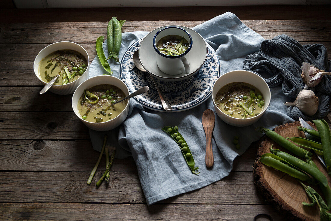 Flat lay of bowls with green pea and coconut cream soup on wooden table with pea pods and garlic in composition