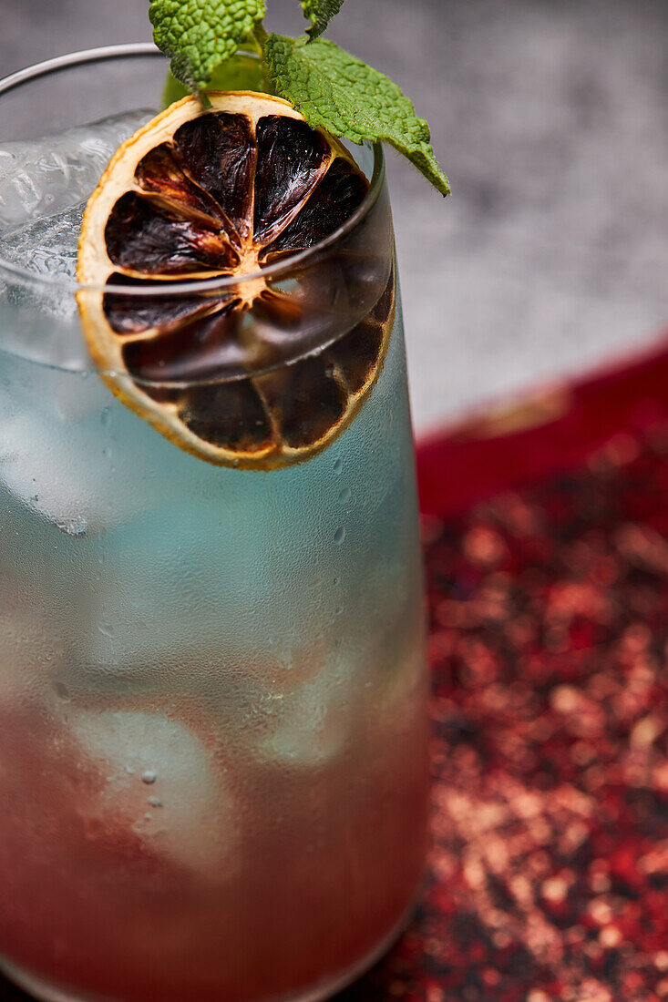 Glass of rainbow paradise colorful cocktail garnished with slice of orange and mint leaf on old book board on table